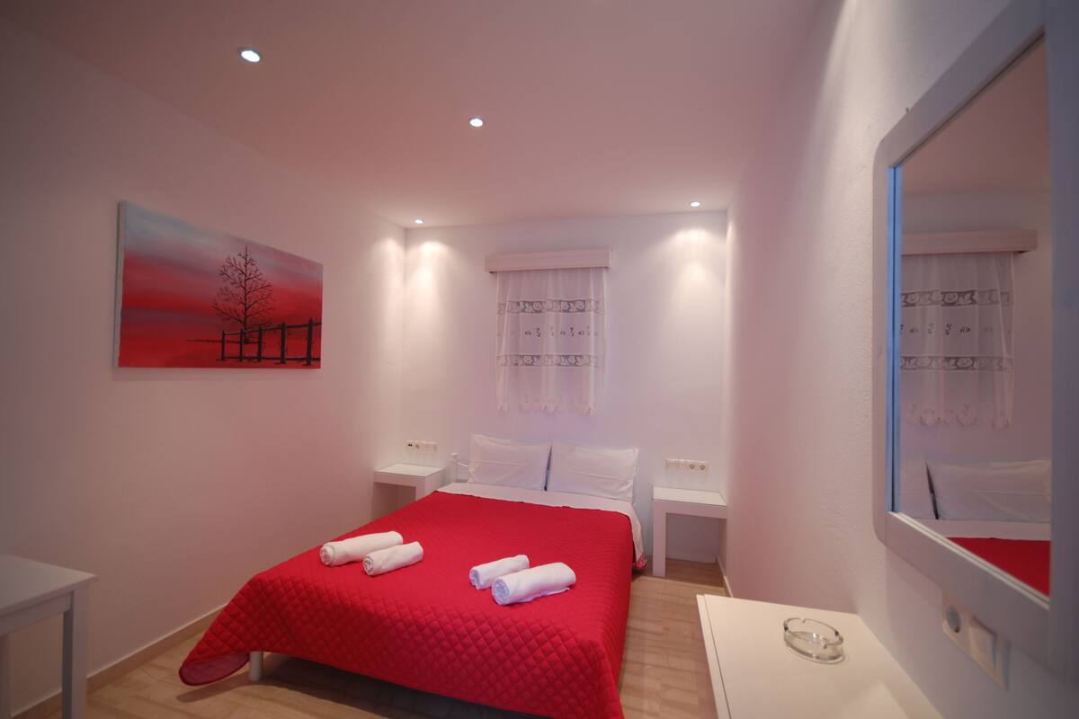 A hotel room with a red bedspread, white pillows, a bedside table, a nightstand, a painting on the wall, and a mirror.
