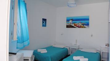 Une chambre d’hôtel avec deux lits, un tableau représentant un paysage côtier, un rideau bleu et une petite table avec une lampe.