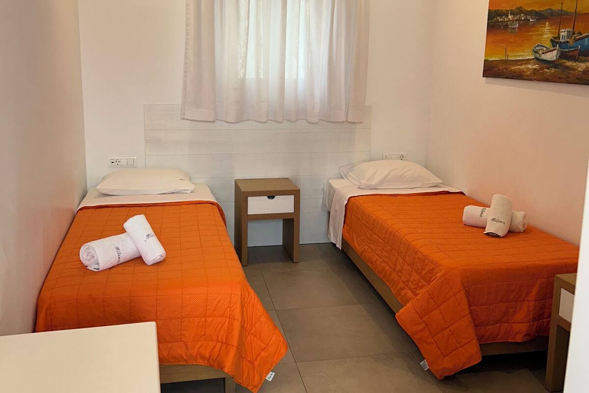 A hotel room with two beds, a small table, a painting on the wall, and a window with curtains.