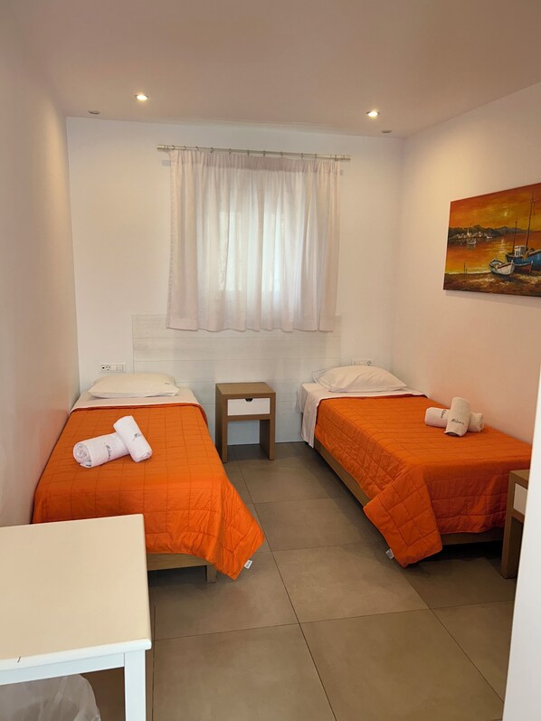 A hotel room with two beds, a small table, a painting on the wall, and a window with curtains. - Milena Hotel (Mykonos)
