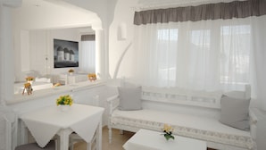 Una habitación luminosa y blanca con un ventanal grande, un sofá blanco, una mesita con un florero y un cuadro en la pared.