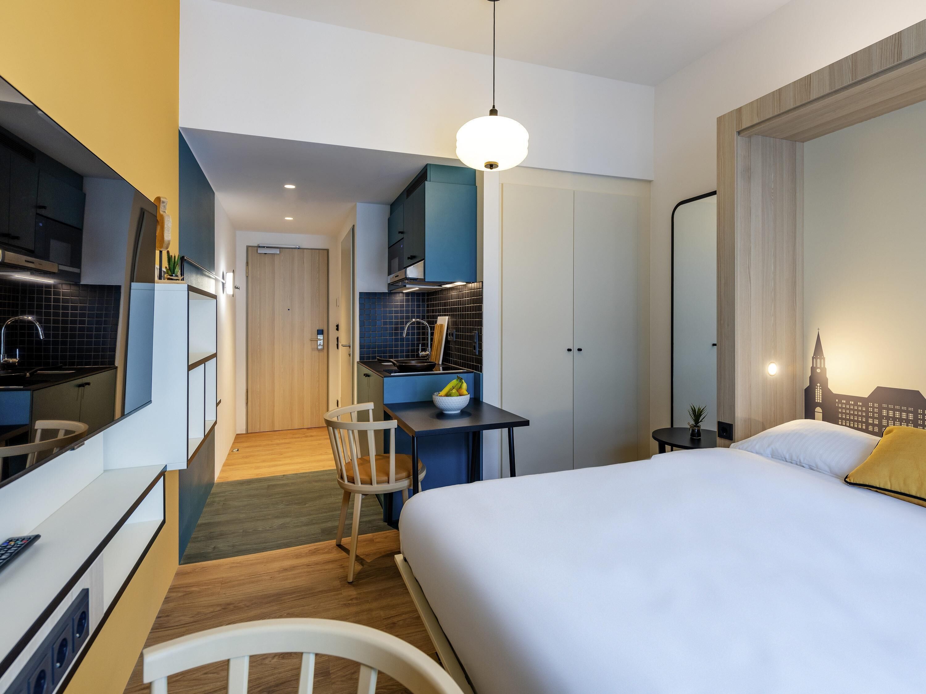 A modern hotel room with a bed, a desk, a chair, a small kitchen area, and a large window.