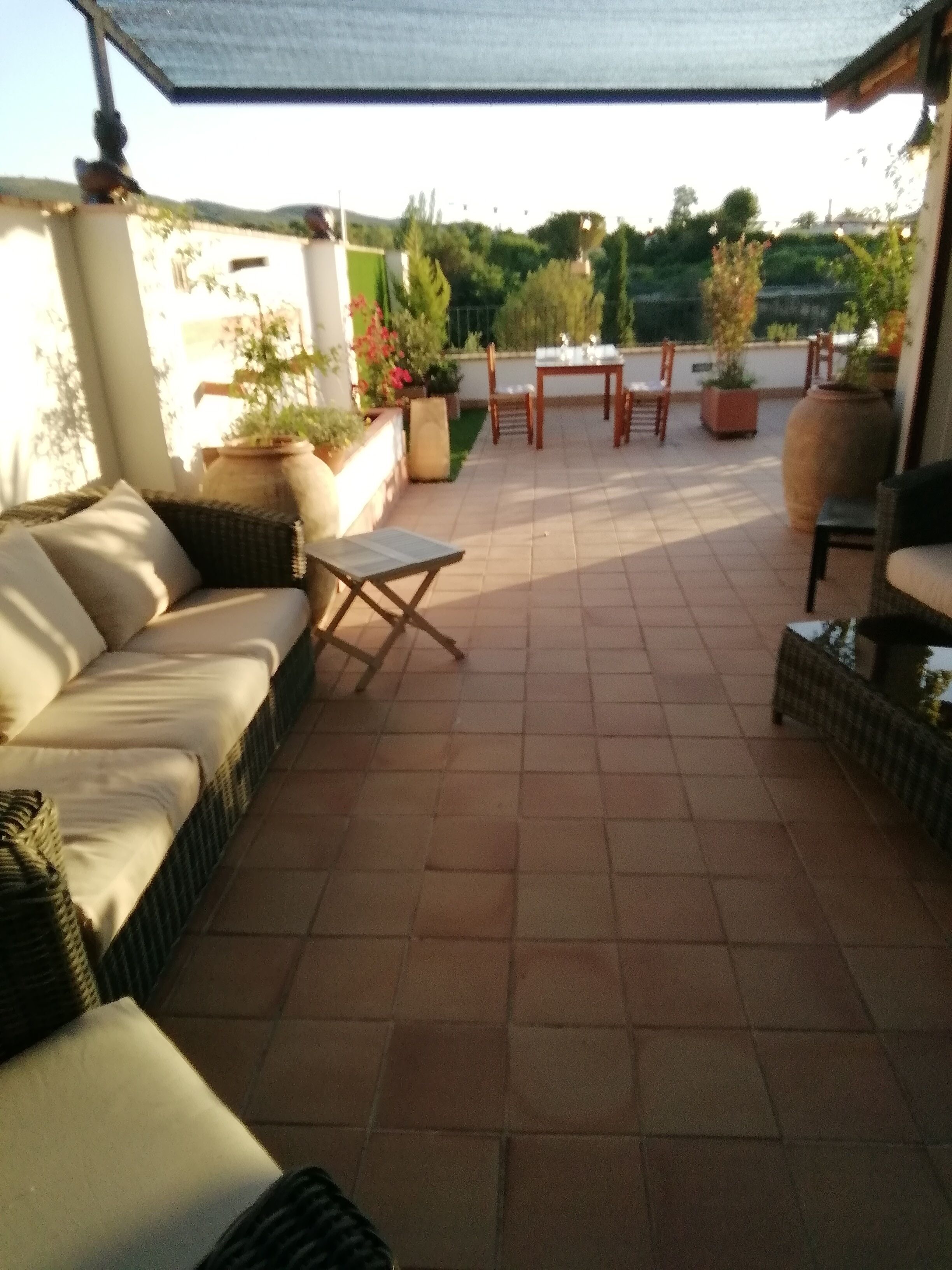A patio with a sofa, a small table, and chairs. There are potted plants and a view of greenery in the background.