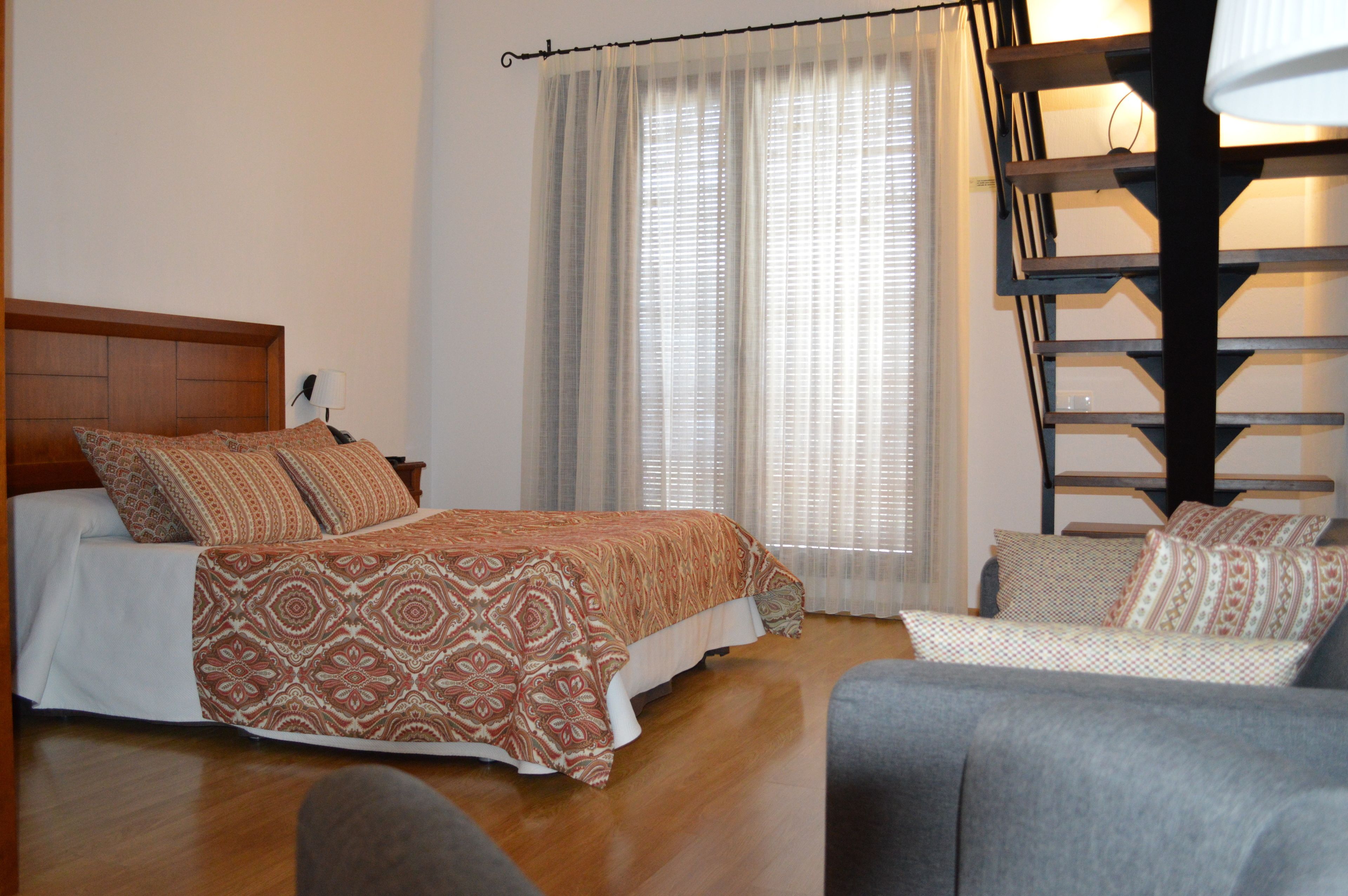 Un dormitorio con cama, sofá, cabecera de madera, ventana con cortinas y una escalera.