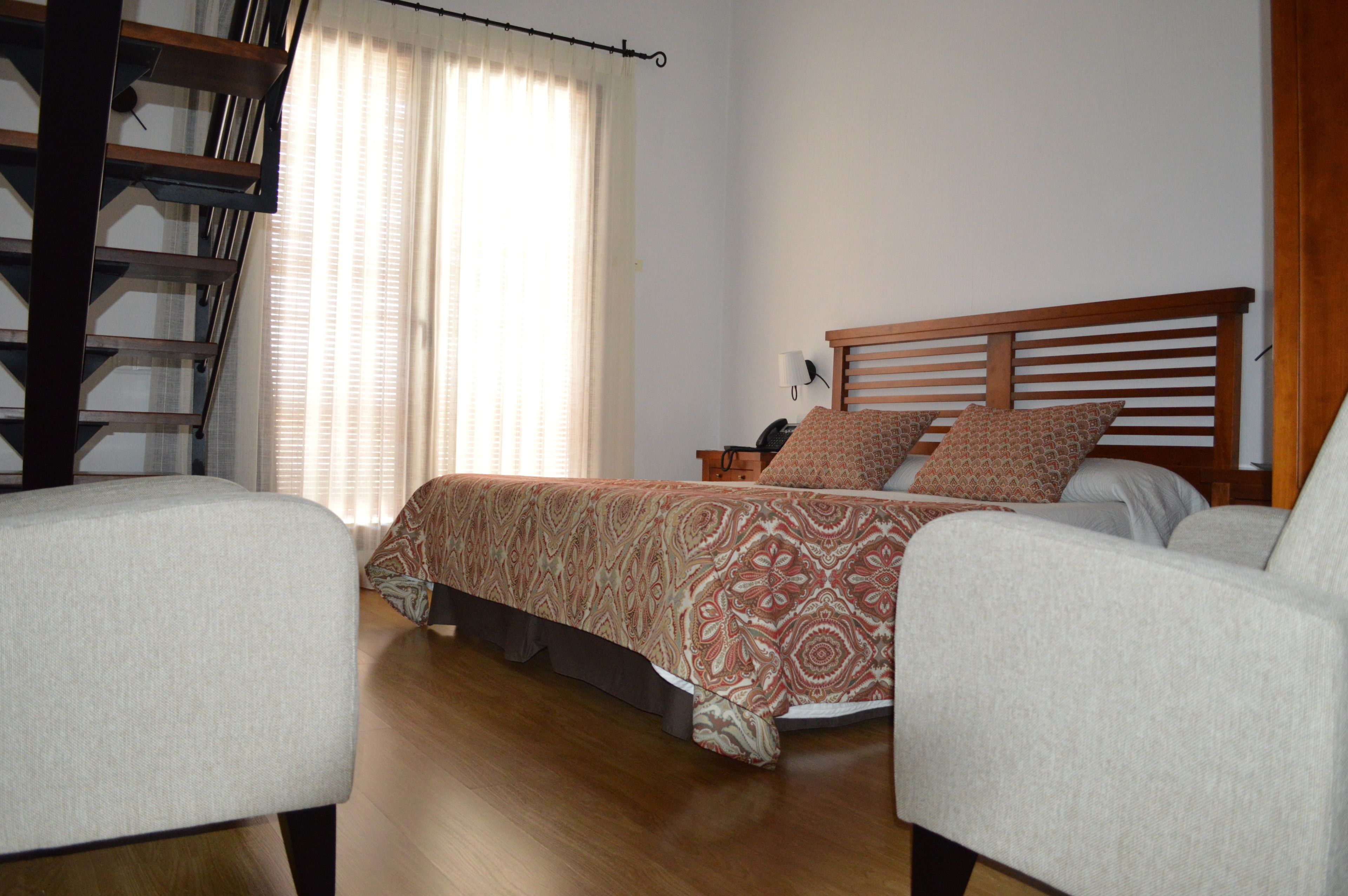 A bedroom with a bed, two chairs, a nightstand, and a window with curtains.