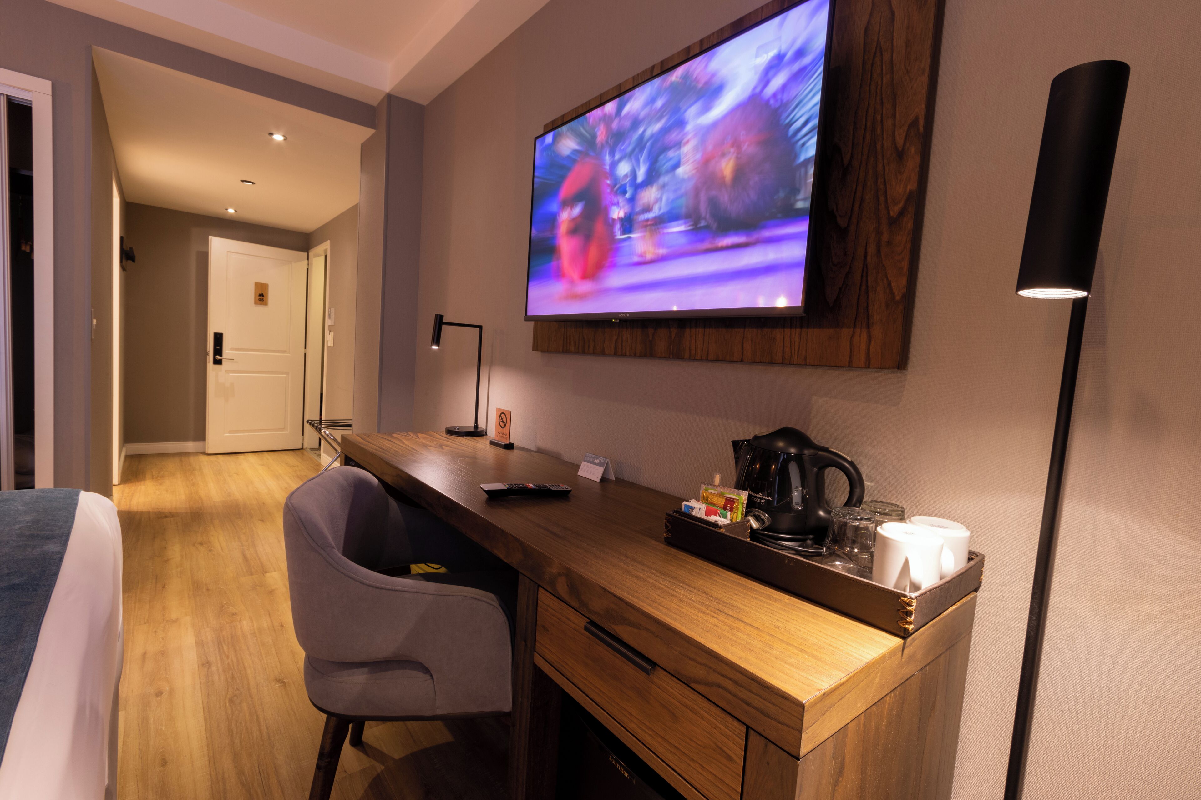 A hotel room with a wooden desk, a television mounted on the wall, a chair, a coffee maker, and a bed.