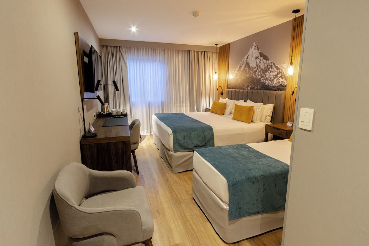 A hotel room with two beds, a desk, a chair, a TV, and a mountain mural on the wall.
