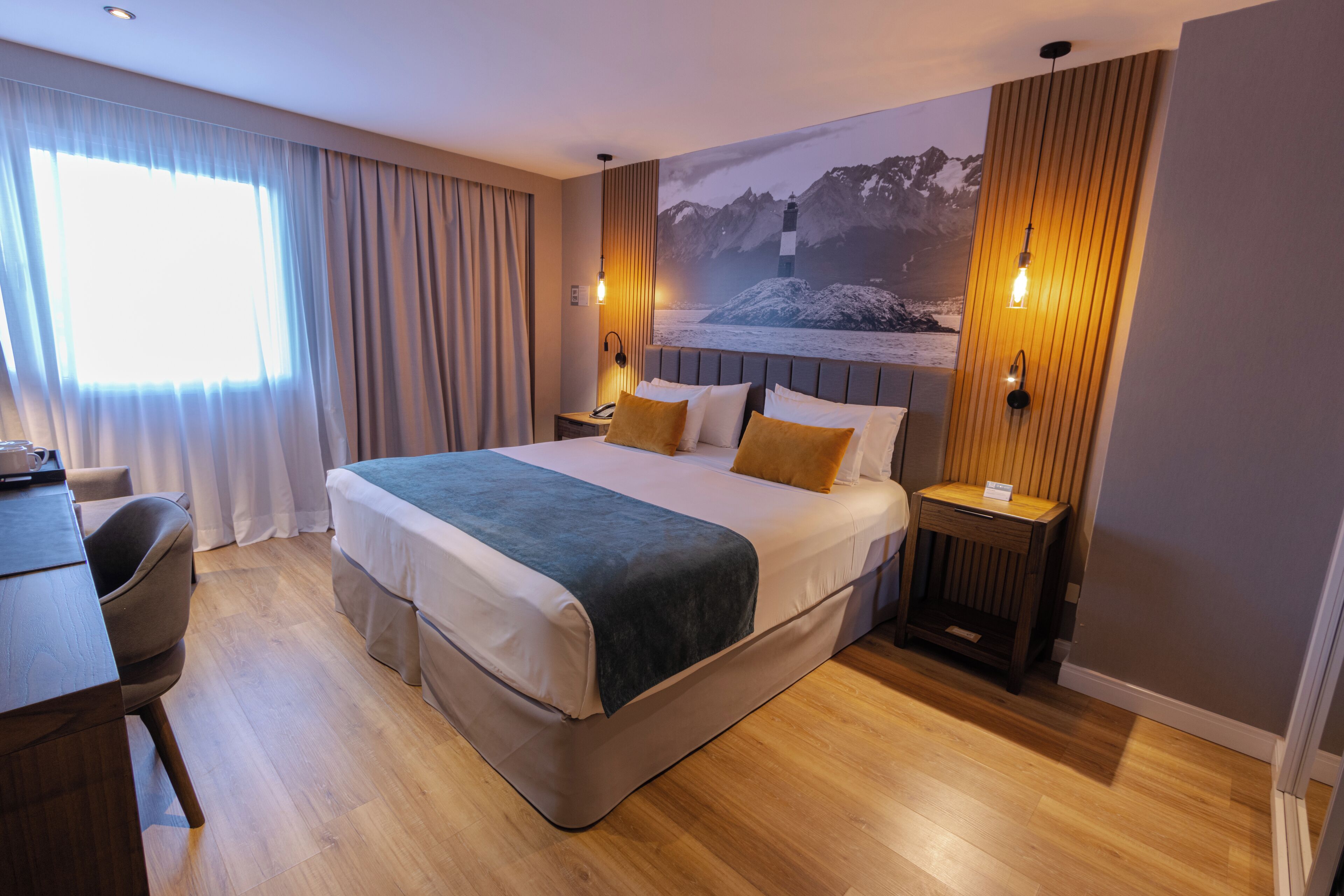 A modern hotel room with a large bed, a desk, and a chair. There is a window with curtains, a wall-mounted lamp, and a painting of a lighthouse.