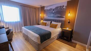 A modern hotel room with a large bed, a desk, and a chair. There is a window with curtains, a wall-mounted lamp, and a painting of a lighthouse.