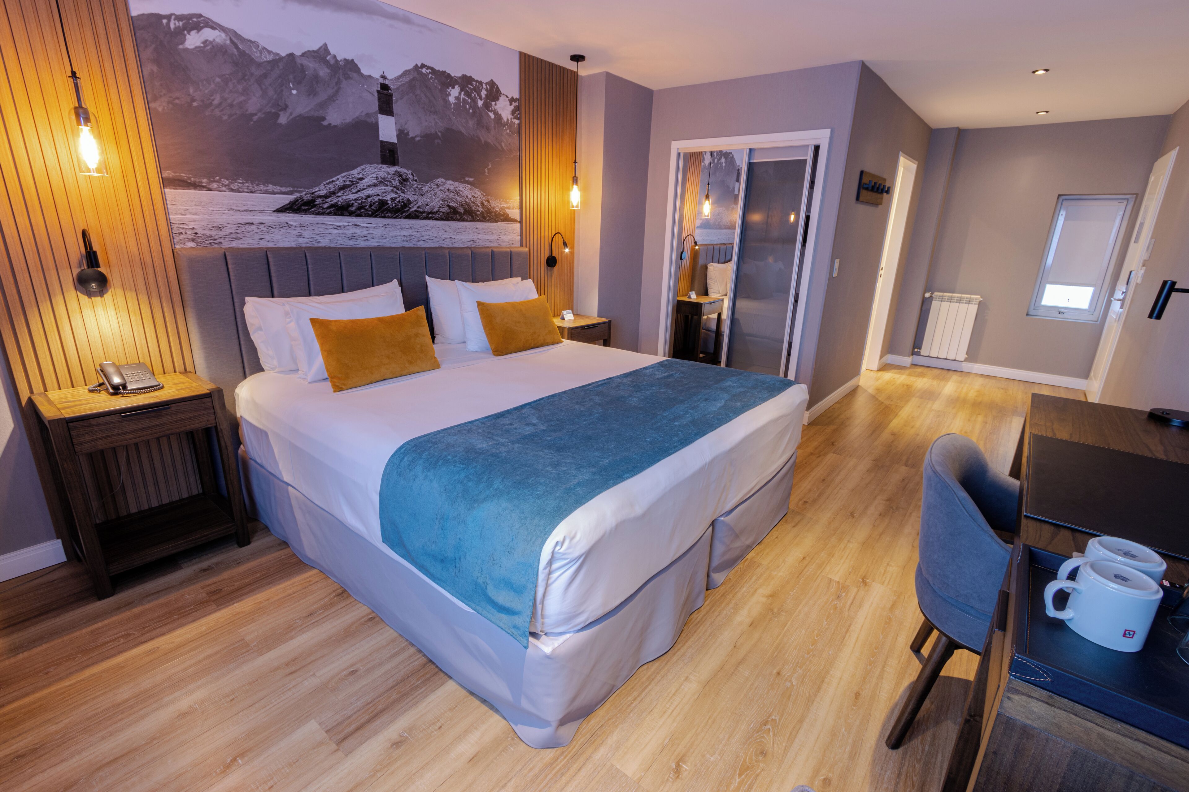 A hotel room with a bed, bedside table, lamp, and a view of mountains and a lighthouse.