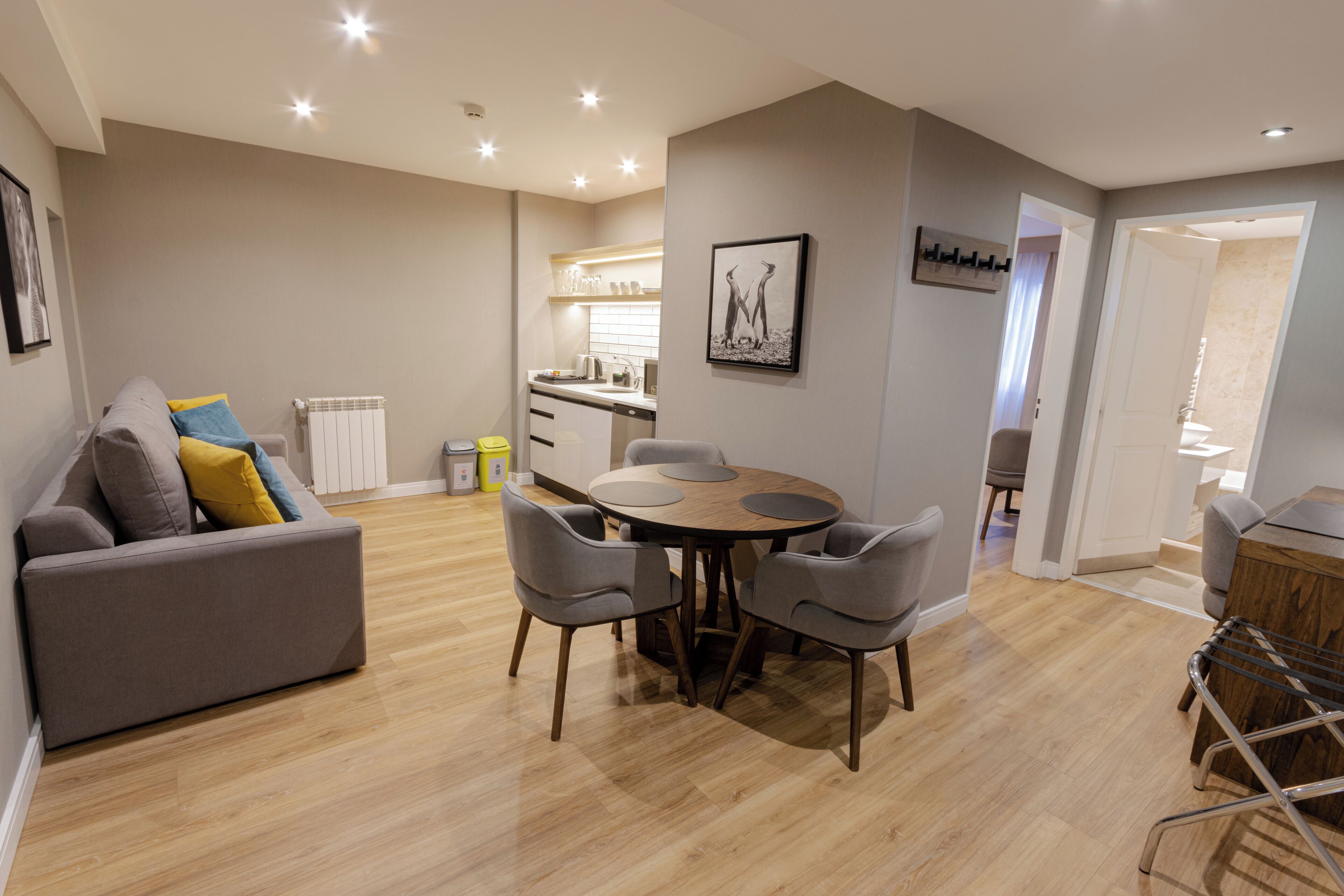 Una sala de estar moderna con un sofá gris, una mesa de comedor redonda con sillas, una cocina y un pasillo que lleva a un baño.
