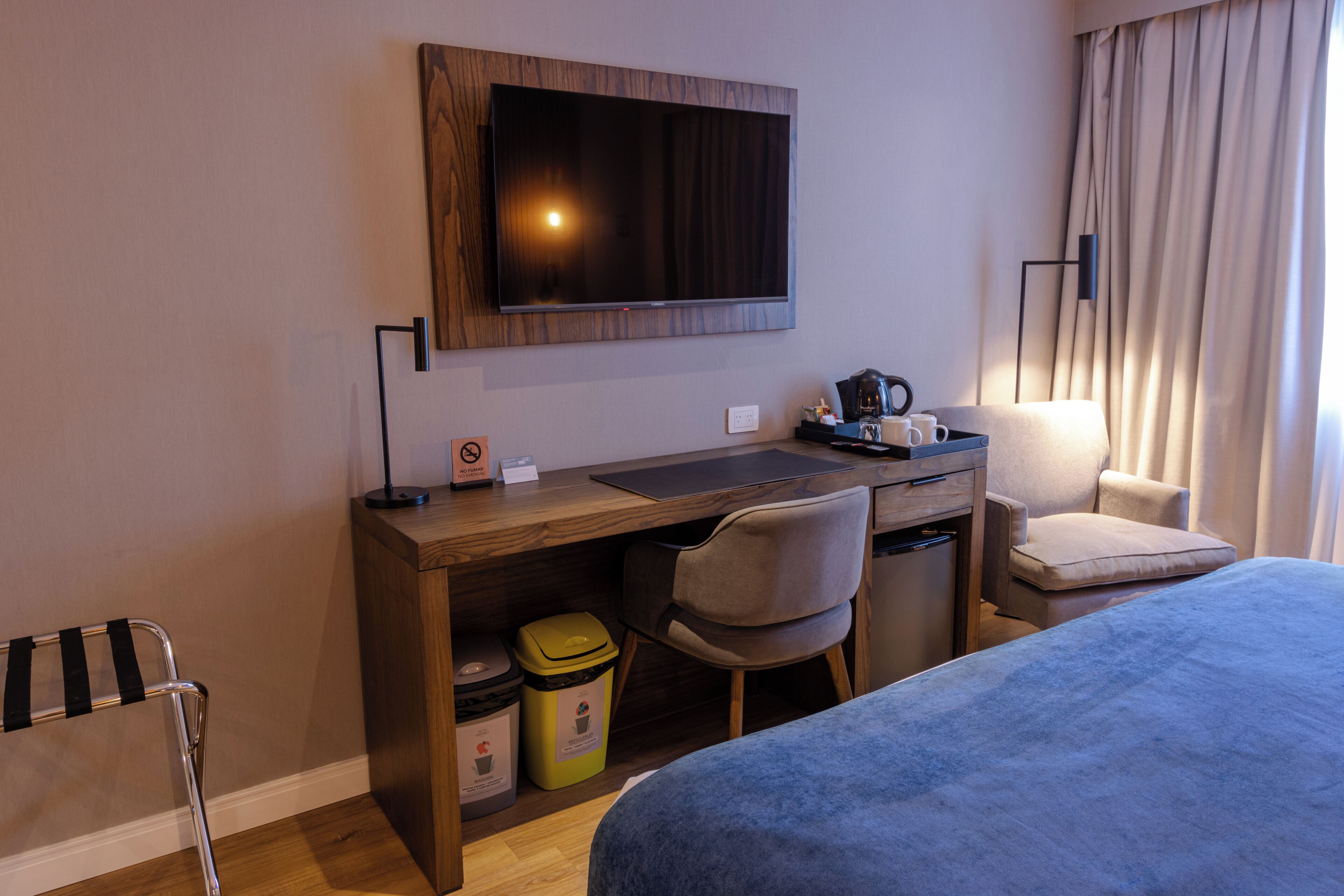 A hotel room with a bed, desk, chair, TV, and a trash bin.
