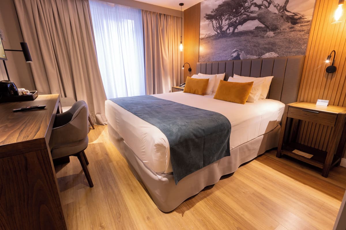 A hotel room with a large bed, a desk, and a chair. There is a window with curtains, a wall-mounted lamp, and a painting on the wall.