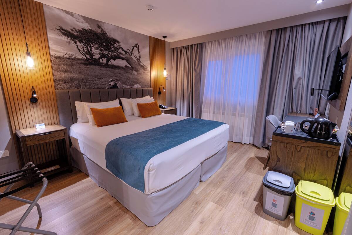 A hotel room with a large bed, wooden paneling, a painting on the wall, a desk with a kettle, and two trash bins.