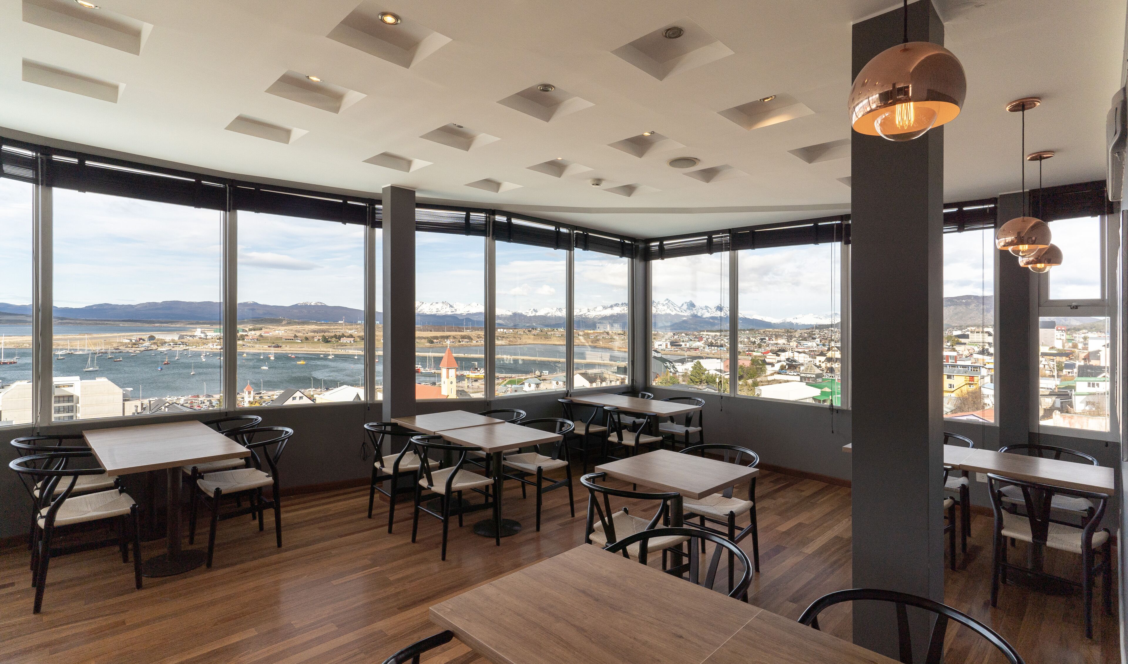 A restaurant with large windows offering a view of a harbor and mountains. Tables and chairs are arranged for dining.