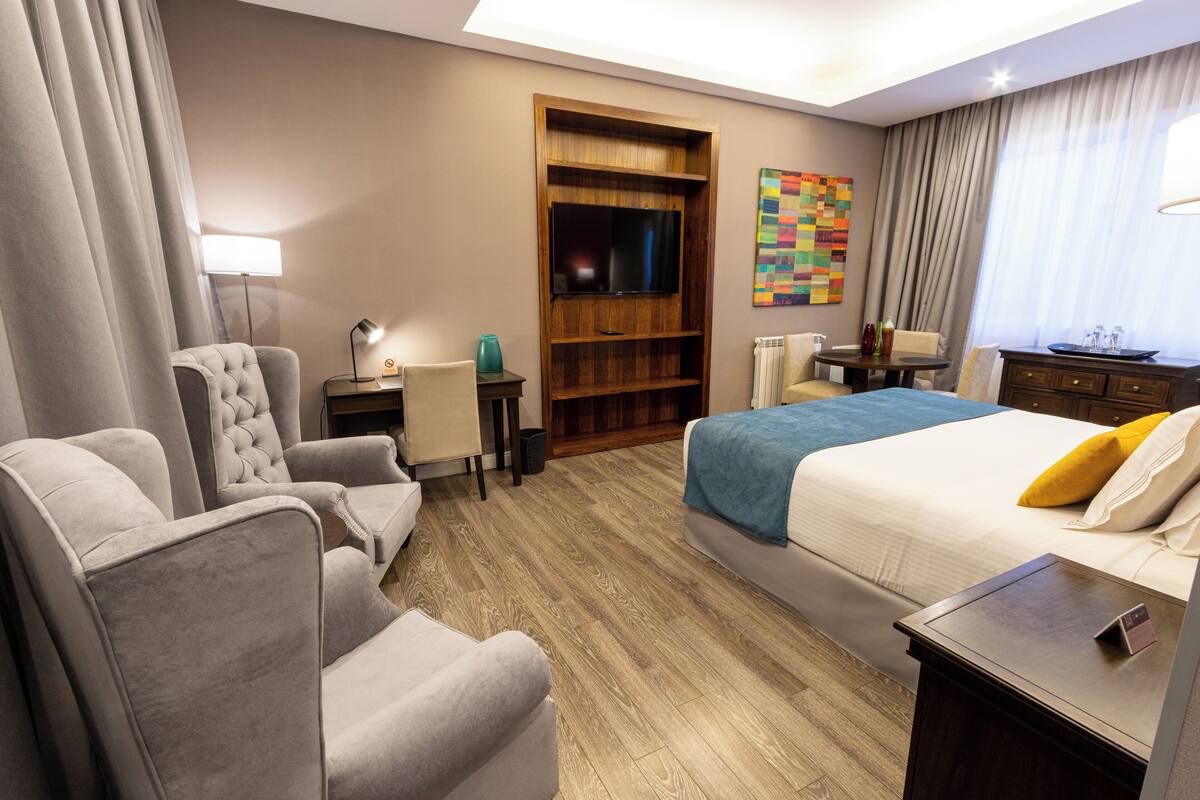 A hotel room with a large bed, two armchairs, a desk, a TV, and a wooden bookshelf.