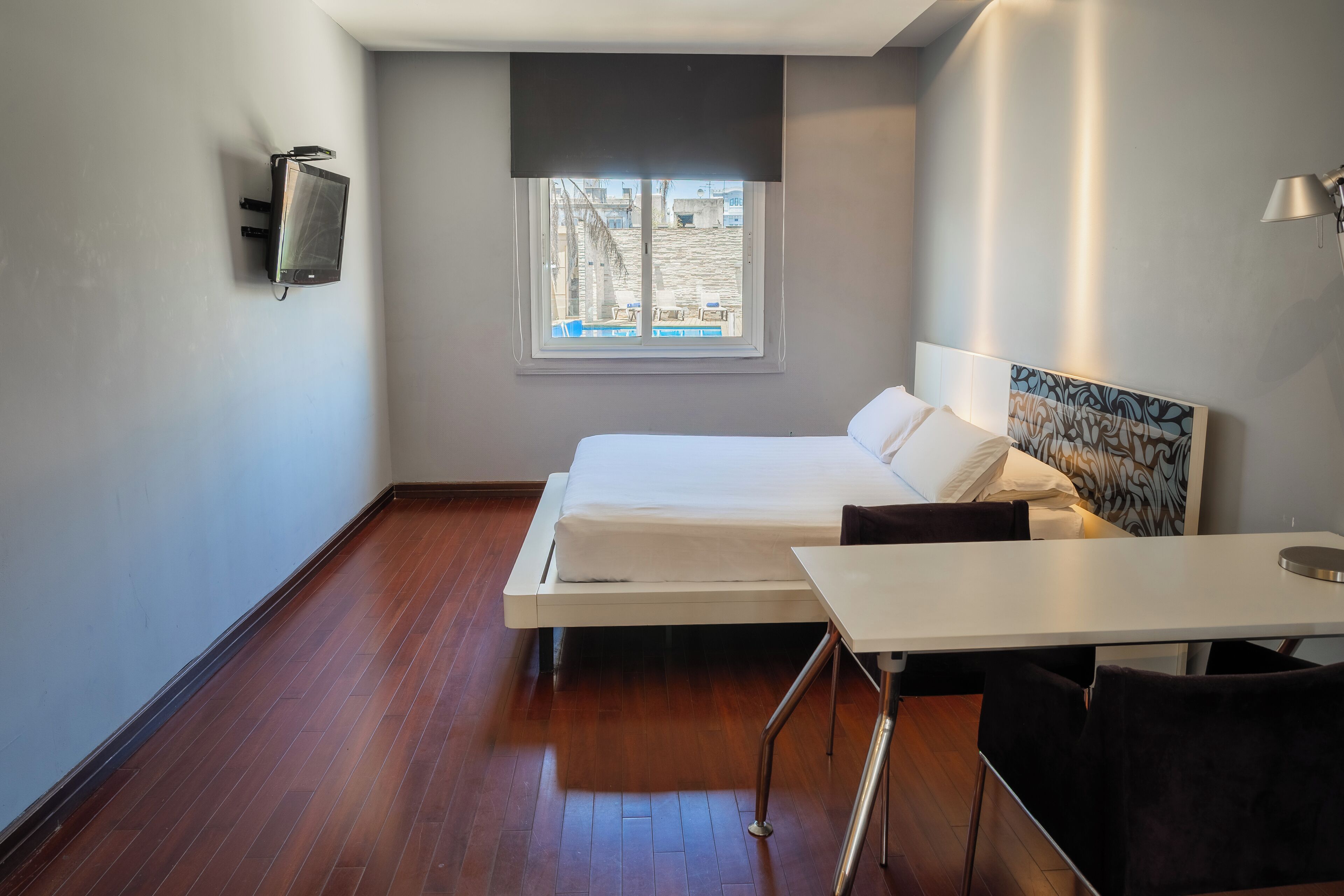 A modern hotel room with a bed, a small table, a chair, a wall-mounted TV, and a window with a view of buildings.