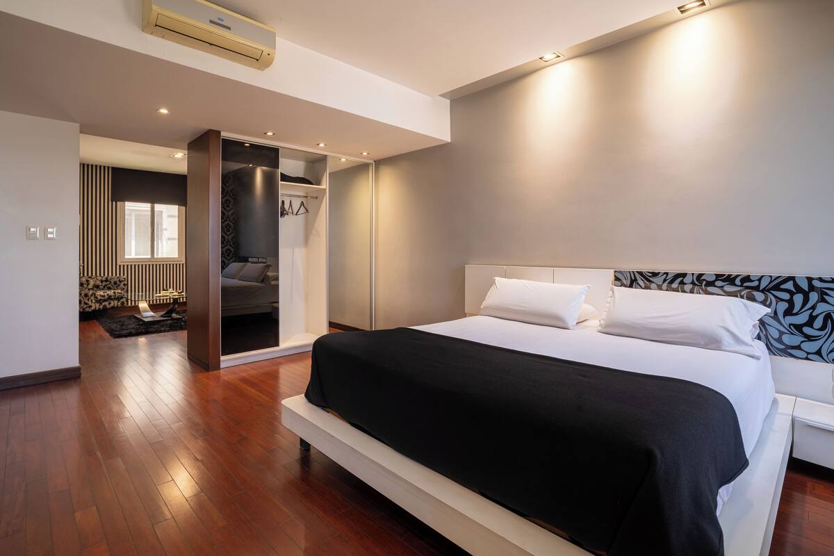 A modern bedroom with a large bed, a walk-in closet, and a view of a living area.