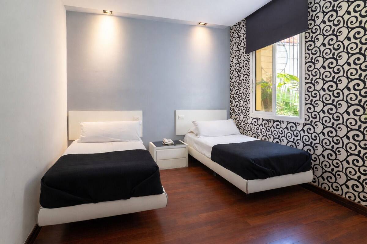 A small, modern room with two beds, a nightstand, and a window with a view of greenery.