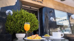 Una mesa de desayuno con platos de comida, un vaso de jugo de naranja y una taza blanca, ubicada frente a un edificio con un letrero que dice "Bon Boutique Hotel".