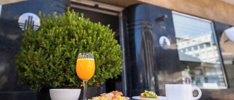 Une table de petit-déjeuner avec des assiettes de nourriture, un verre de jus d’orange et une tasse blanche, placée à l’extérieur d’un bâtiment sur lequel on peut lire l’inscription « Bon Boutique Hotel ».