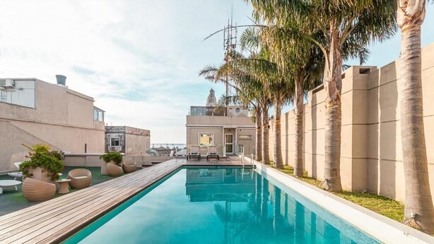 Una piscina rectangular con muelle de madera, rodeada de palmeras y edificios modernos.