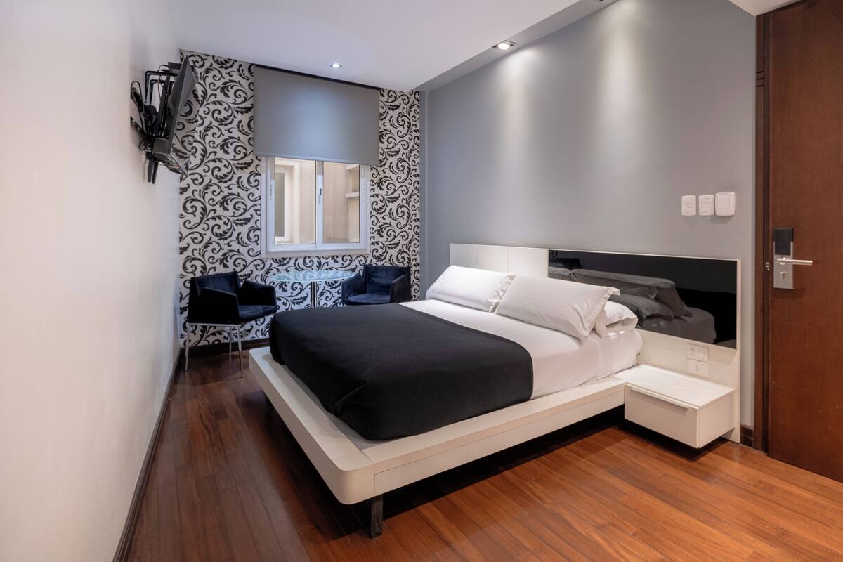 A modern bedroom with a large bed, bedside tables, a built-in TV, and a window with patterned curtains.