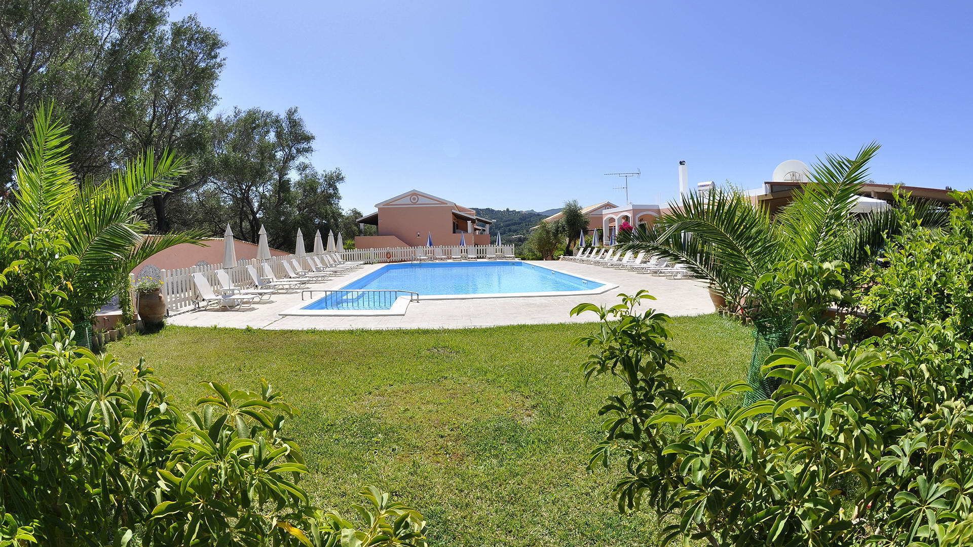 A swimming pool surrounded by lounge chairs and umbrellas, with a white fence and greenery in the foreground.