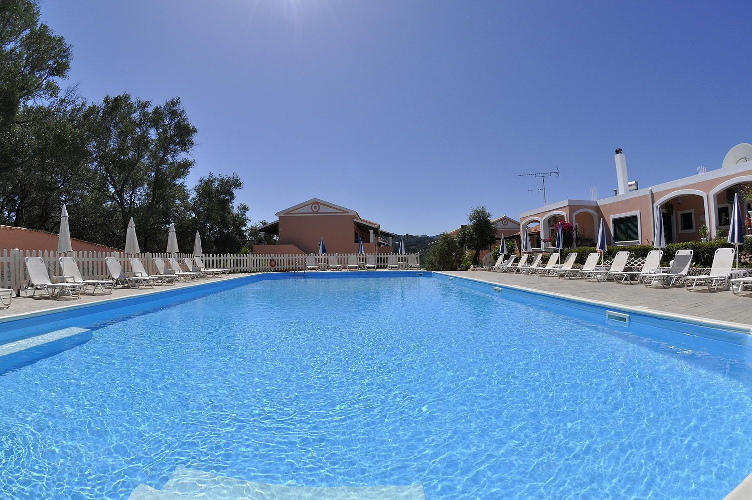 A clear blue swimming pool with lounge chairs and umbrellas, surrounded by a white picket fence and buildings with arched windows.