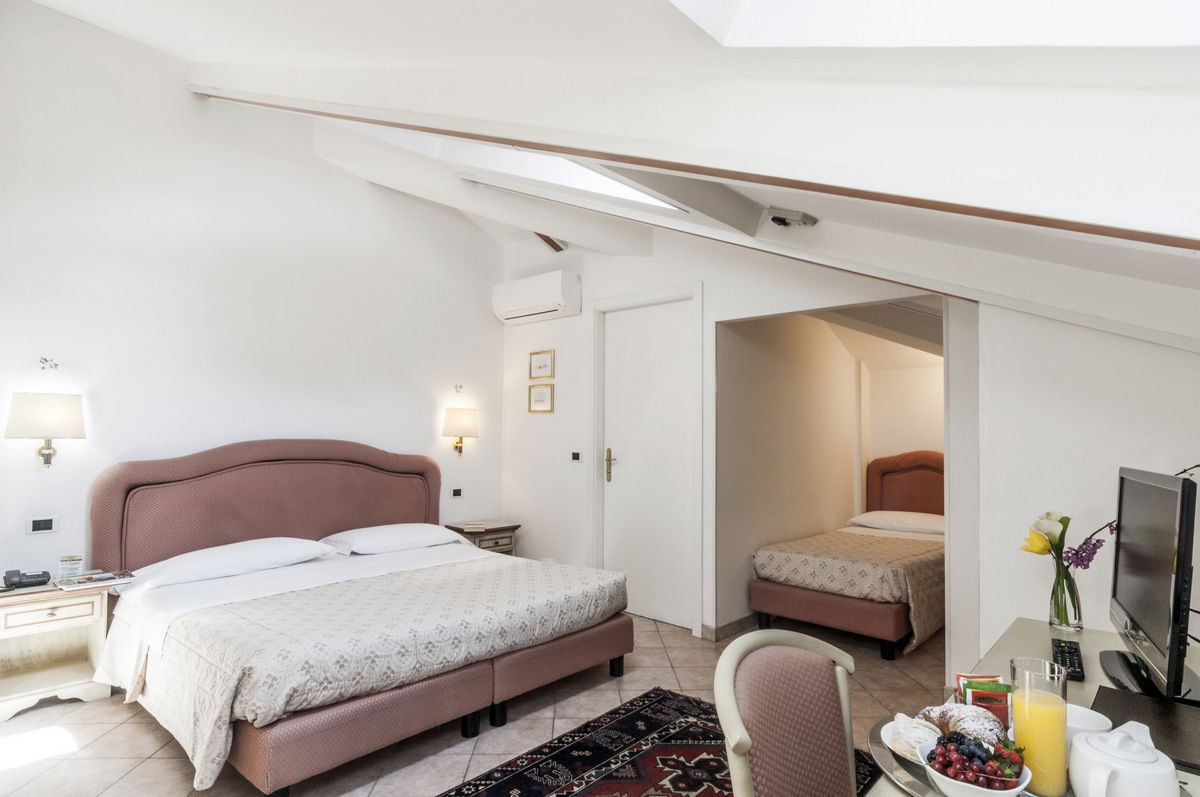 Een hotelkamer met een groot bed, een kleiner bed, een bureau met een televisie en een ontbijttafel met fruit en sap.