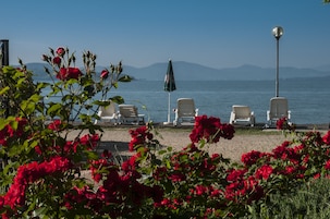 A beachside seating area with white chairs and a green umbrella, overlooking a calm sea and distant mountains.