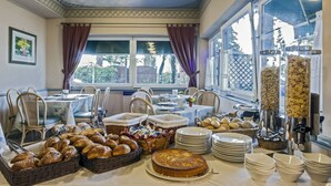 Un bufé de desayuno con diferentes panes, bollería y un pastel, ubicado en una sala con ventanas amplias y un techo con diseño geométrico.