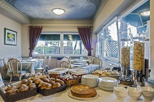 A breakfast buffet with various breads, pastries, and a cake, set in a room with large windows and a ceiling with a geometric pattern.