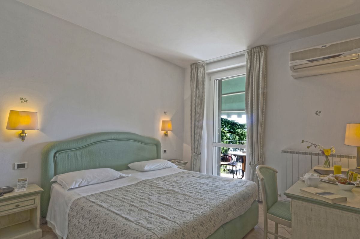 A hotel room with a bed, bedside lamps, a desk with a chair, and a window with curtains.