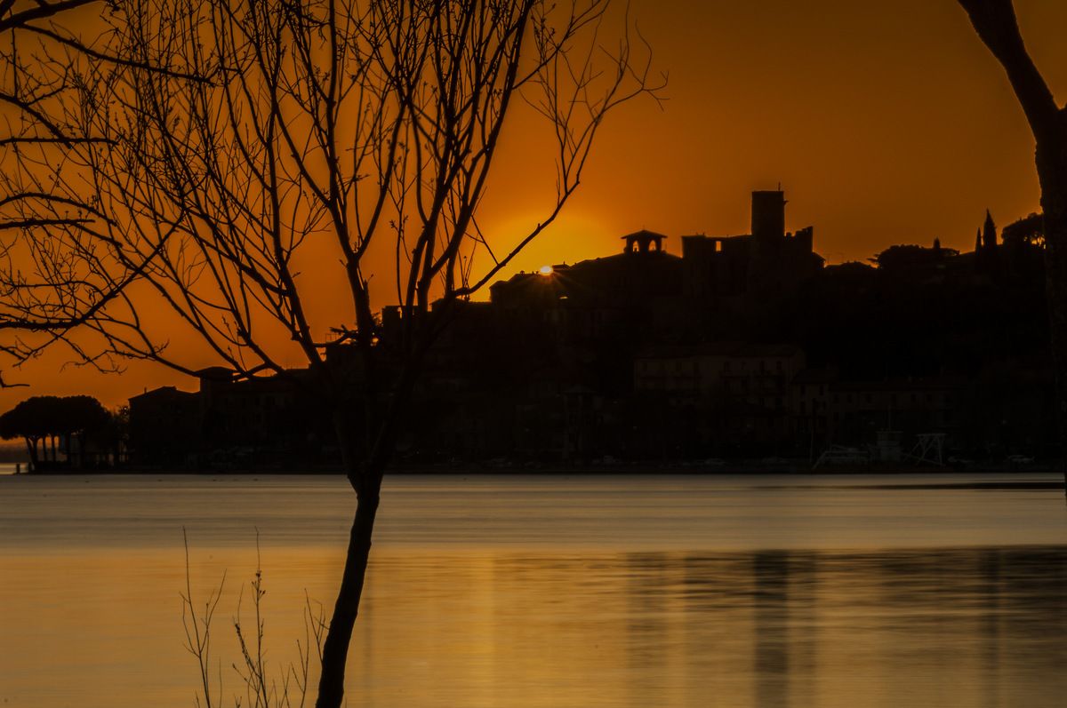 A silhouette of a city skyline at sunset with buildings reflected in calm water.
