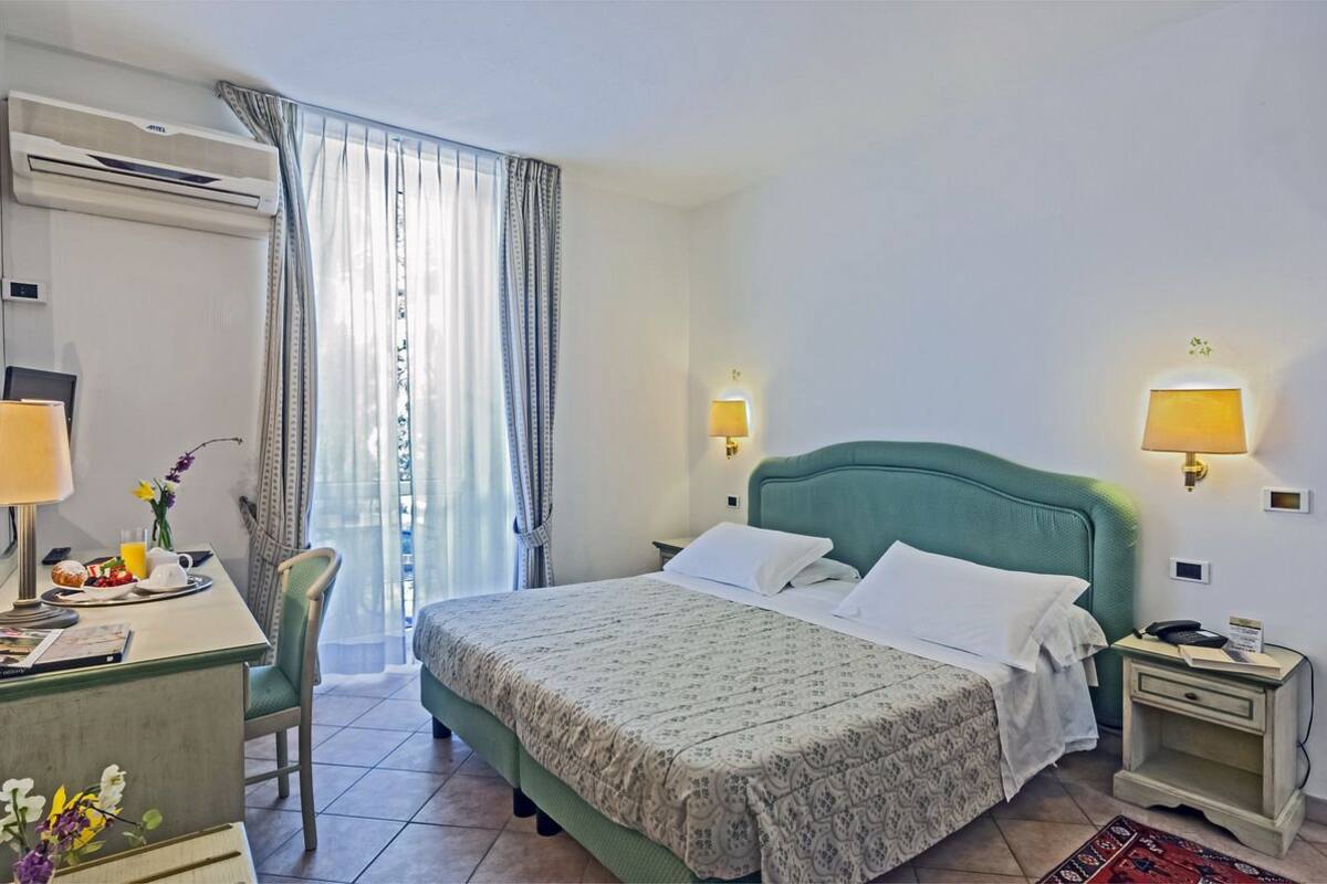 A hotel room with a bed, bedside lamps, a desk with a chair, a window with curtains, and an air conditioning unit.