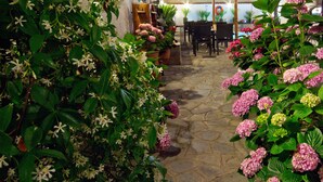 A stone pathway flanked by lush greenery and pink hydrangea bushes, leading to a wooden pergola with tables and chairs.