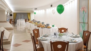 Un área de comedor con mesas redondas preparadas para una comida, sillas y una mesa larga al fondo.