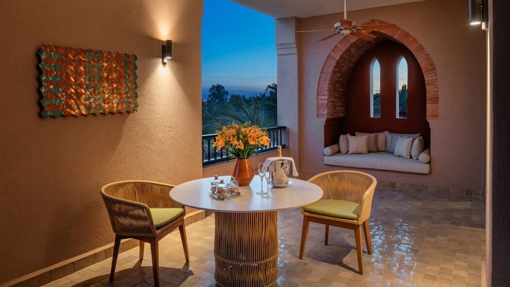 Un comedor con una mesa redonda, dos sillas de mimbre, un florero con flores y vistas al exterior.