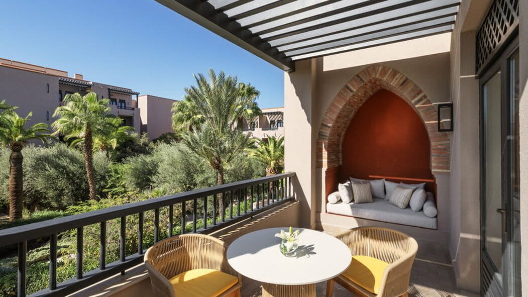 A balcony with wicker chairs and a table, a cushioned seating area with a red arch, and a view of palm trees and buildings.