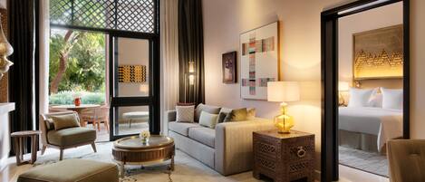 A modern hotel room with a sofa, armchairs, a round coffee table, a side table with a lamp, and a bed with white linens.