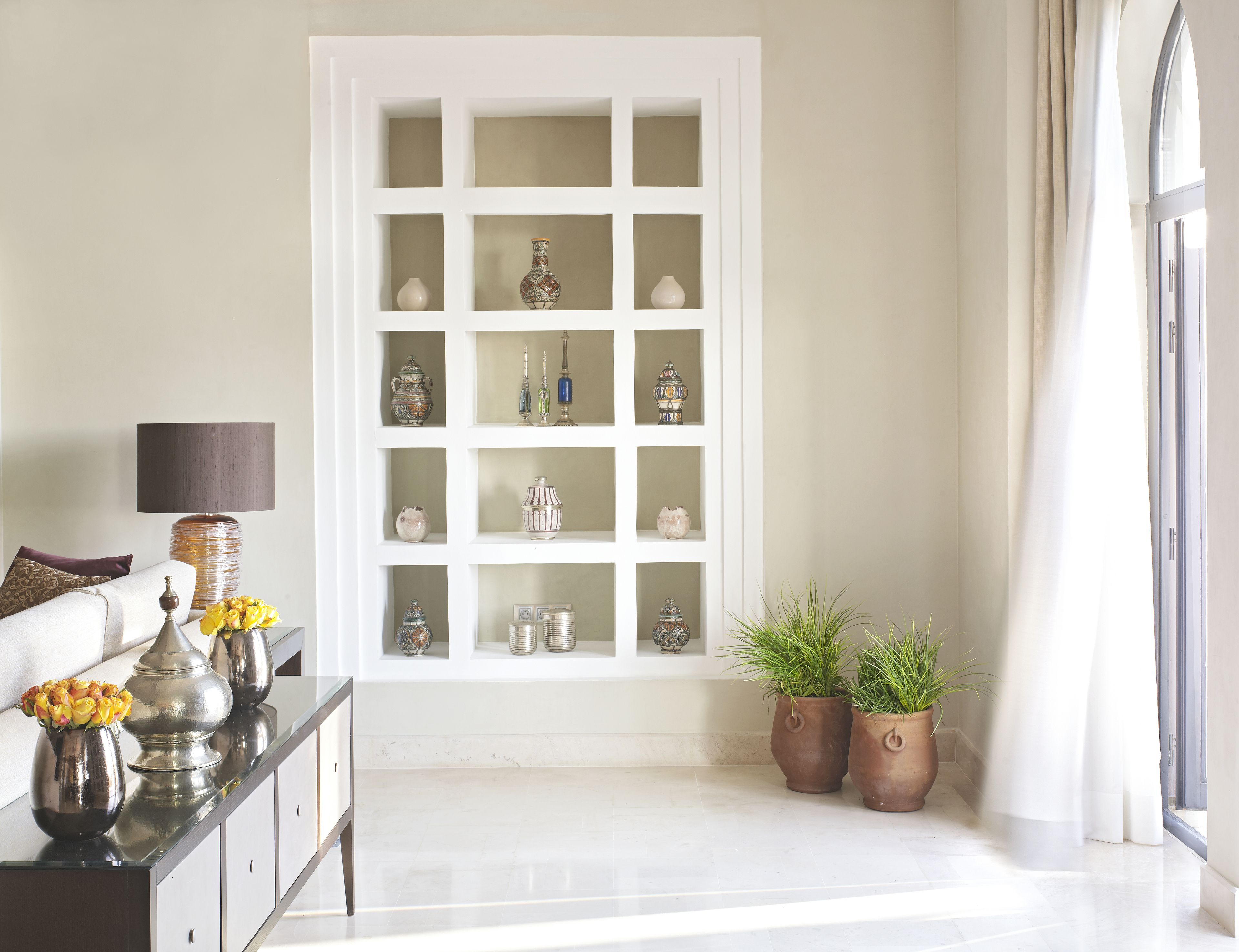 Una habitación con un aparador blanco con puertas de vidrio que exhibe varios objetos decorativos, un sofá con cojines, una lámpara y dos plantas en macetas.