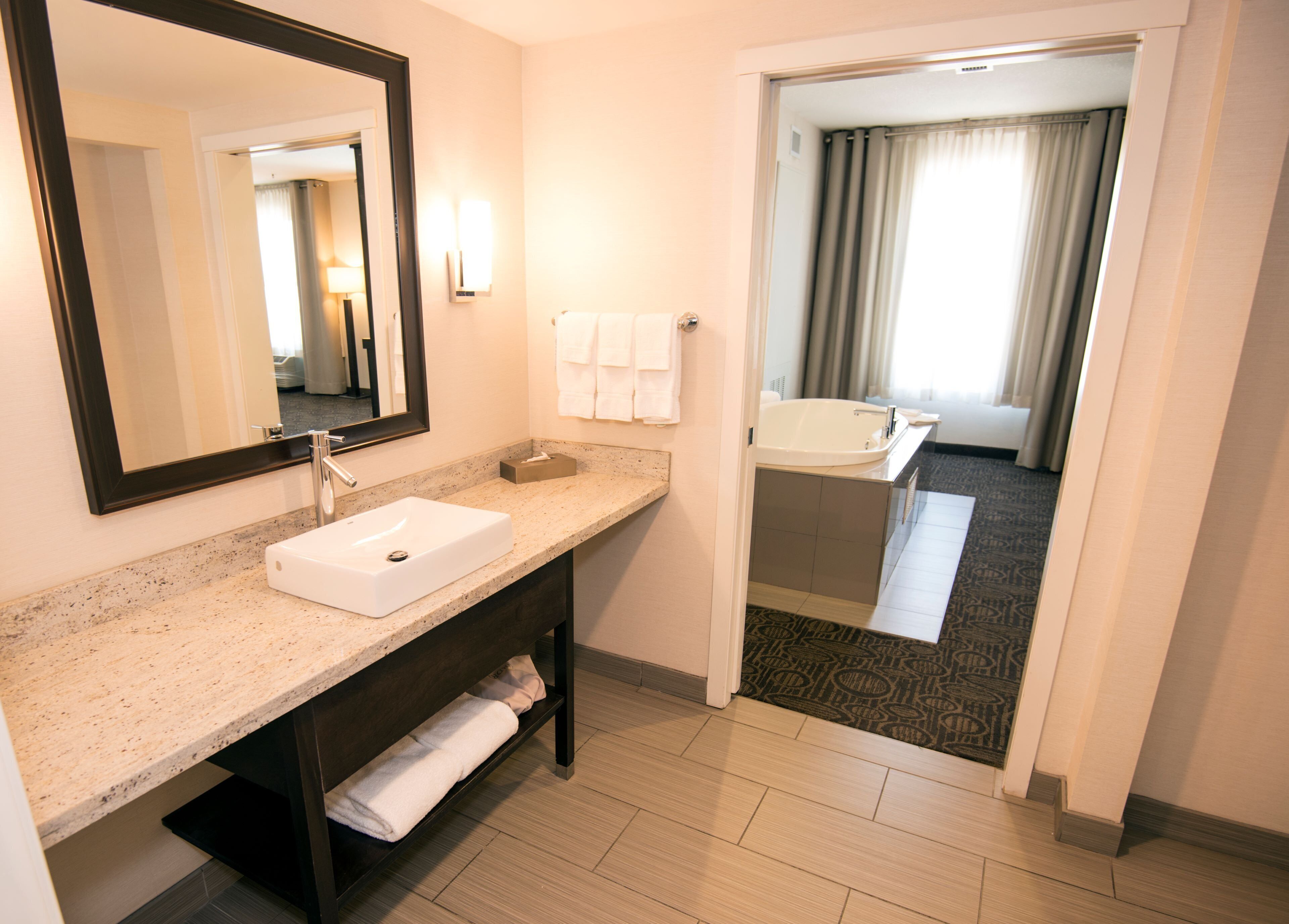 A hotel bathroom with a sink, mirror, towel rack, and a view of a bedroom with a bathtub.