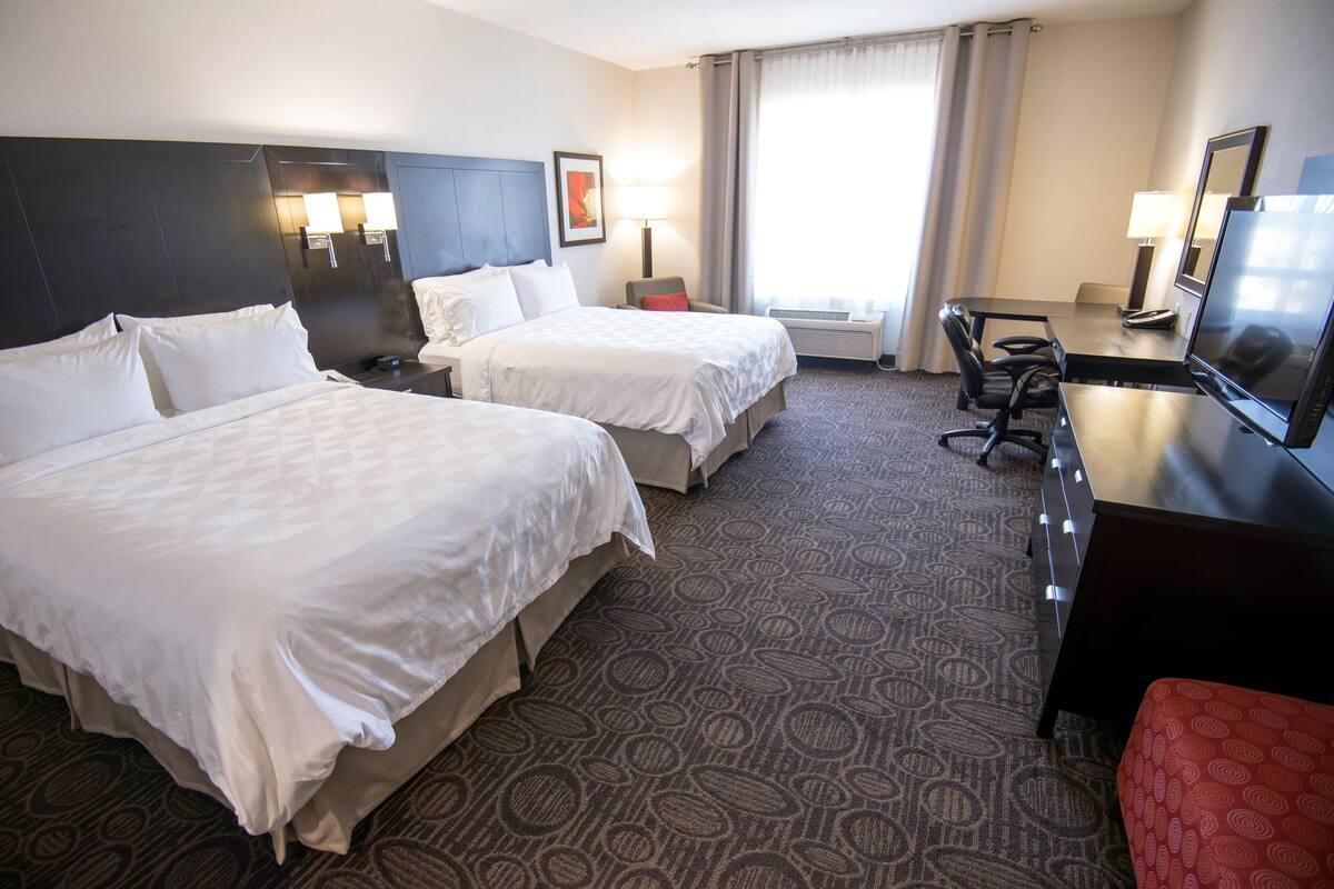 A hotel room with two beds, a desk, a chair, a TV, and a lamp.