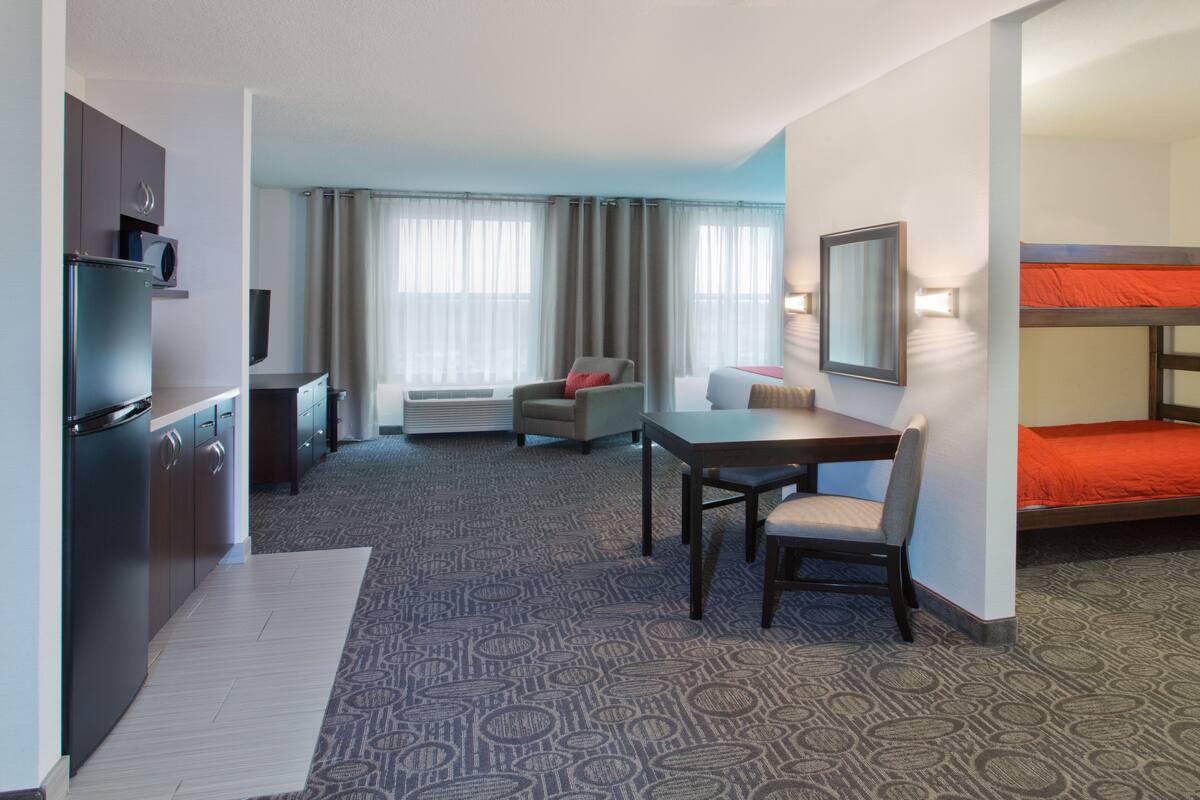 A hotel room with a kitchenette, a dining area with a table and chairs, a single bed, a TV, and a large window with curtains.