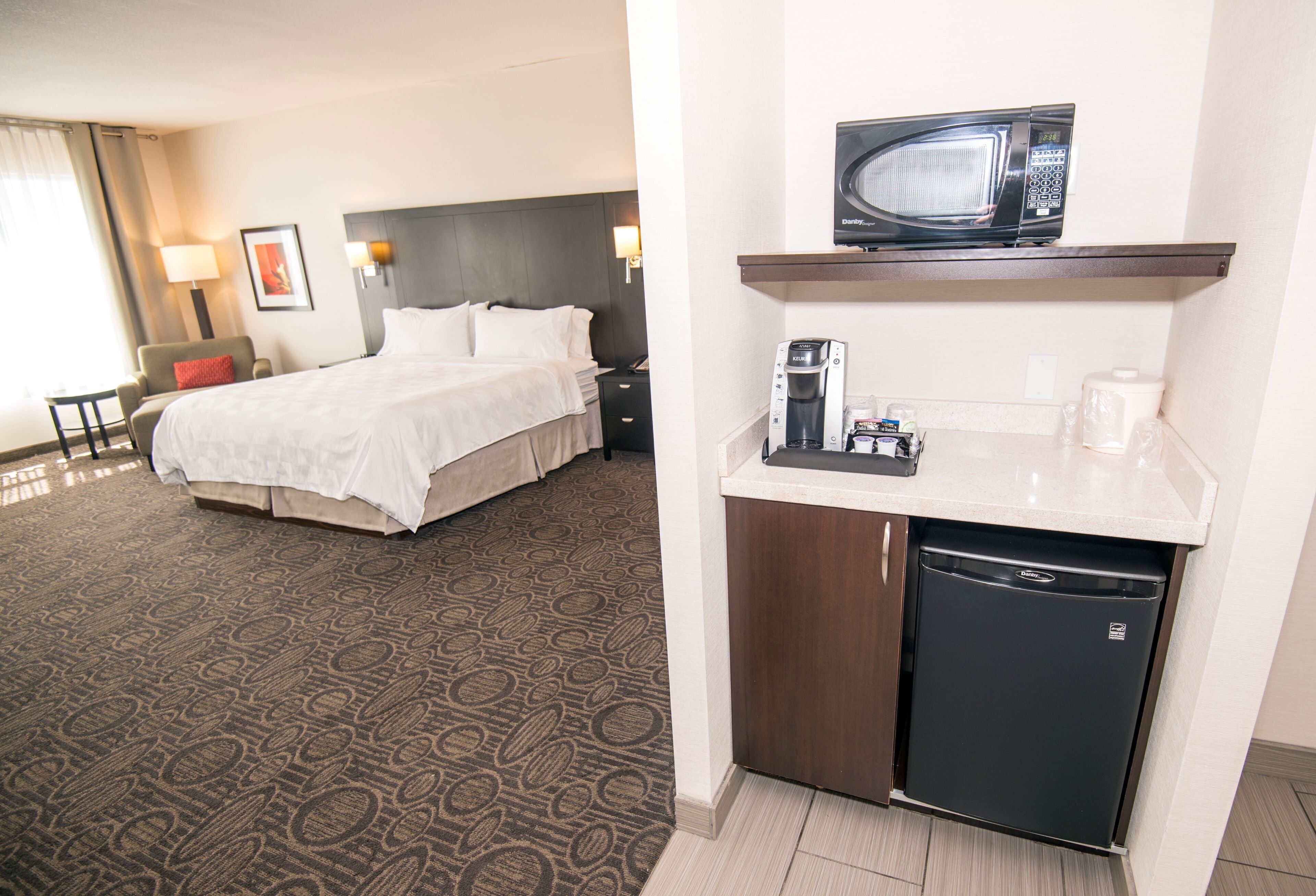 A hotel room with a bed, bedside table, lamp, and a kitchenette with a microwave, coffee maker, and refrigerator.