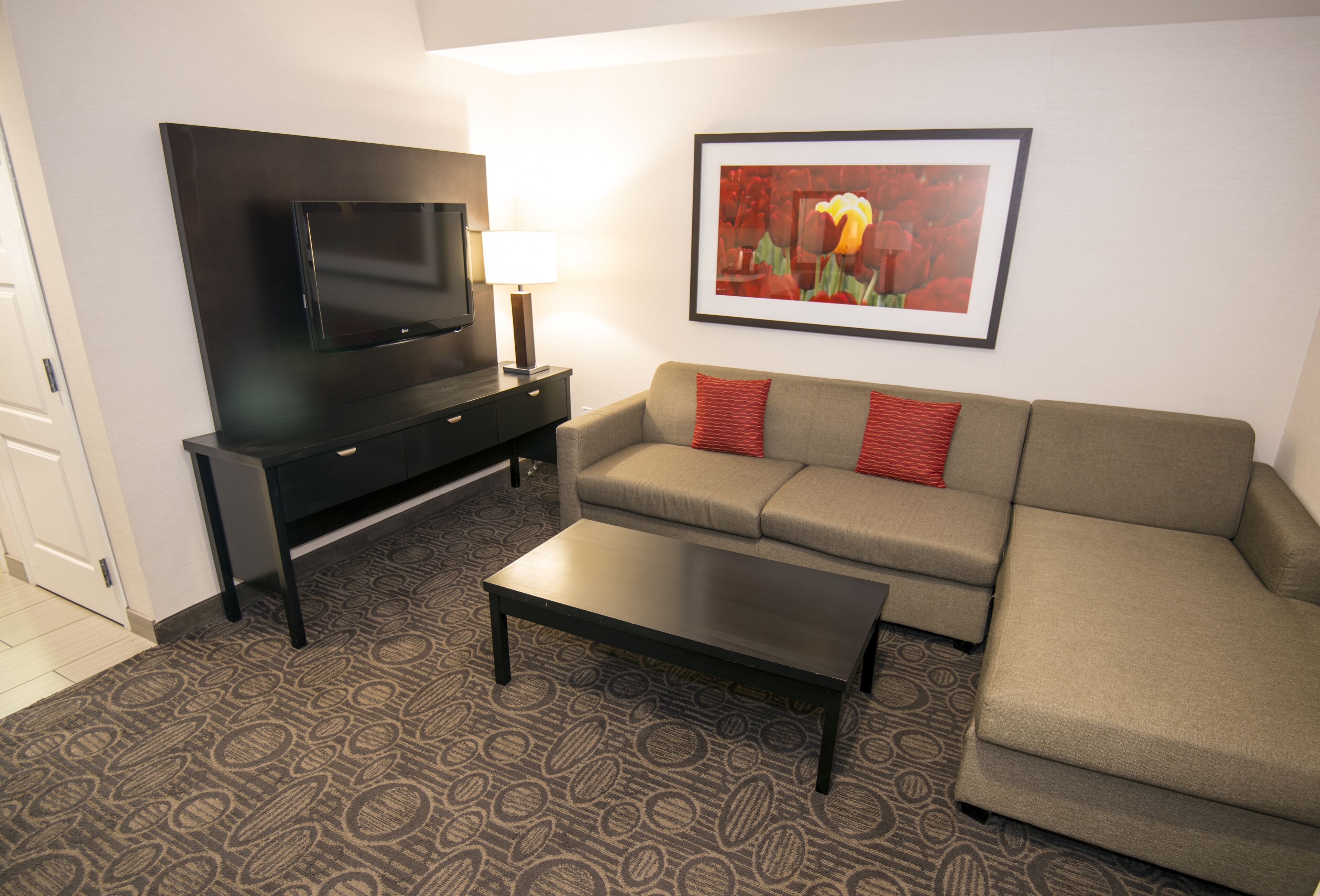 A living room with a flat-screen TV, a sofa with red cushions, a coffee table, and a patterned carpet.