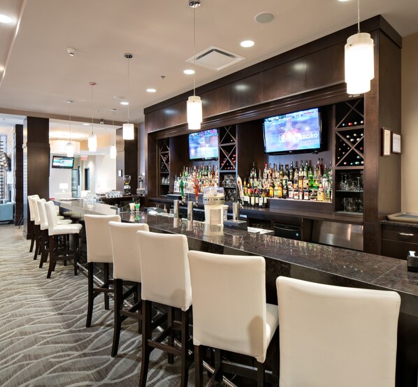 A bar area with a long counter, multiple bar stools, and a television screen displaying a sports event. - Holiday Inn Hotel & Suites Red Deer South by IHG (Red Deer)
