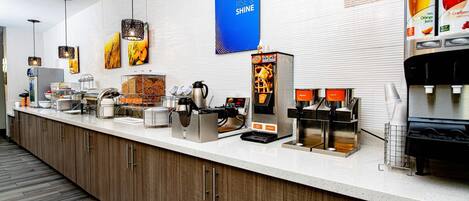 A hotel reception area with a counter, coffee machines, and a wall with a 'RISE & SHINE' sign.