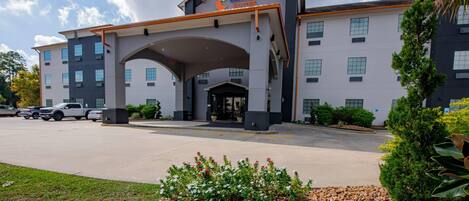 A Comfort Suites hotel with a modern design, featuring a large entrance canopy, multiple windows, and a parking lot with several cars.